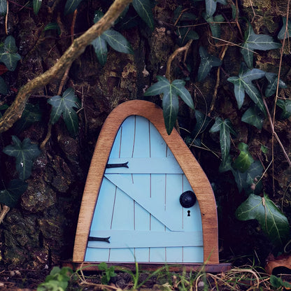 Miniature enchanted fairy garden door set against a backdrop of leaves and twigs, resembling a small wooden house integrated into a natural tree setting, surrounded by grass.