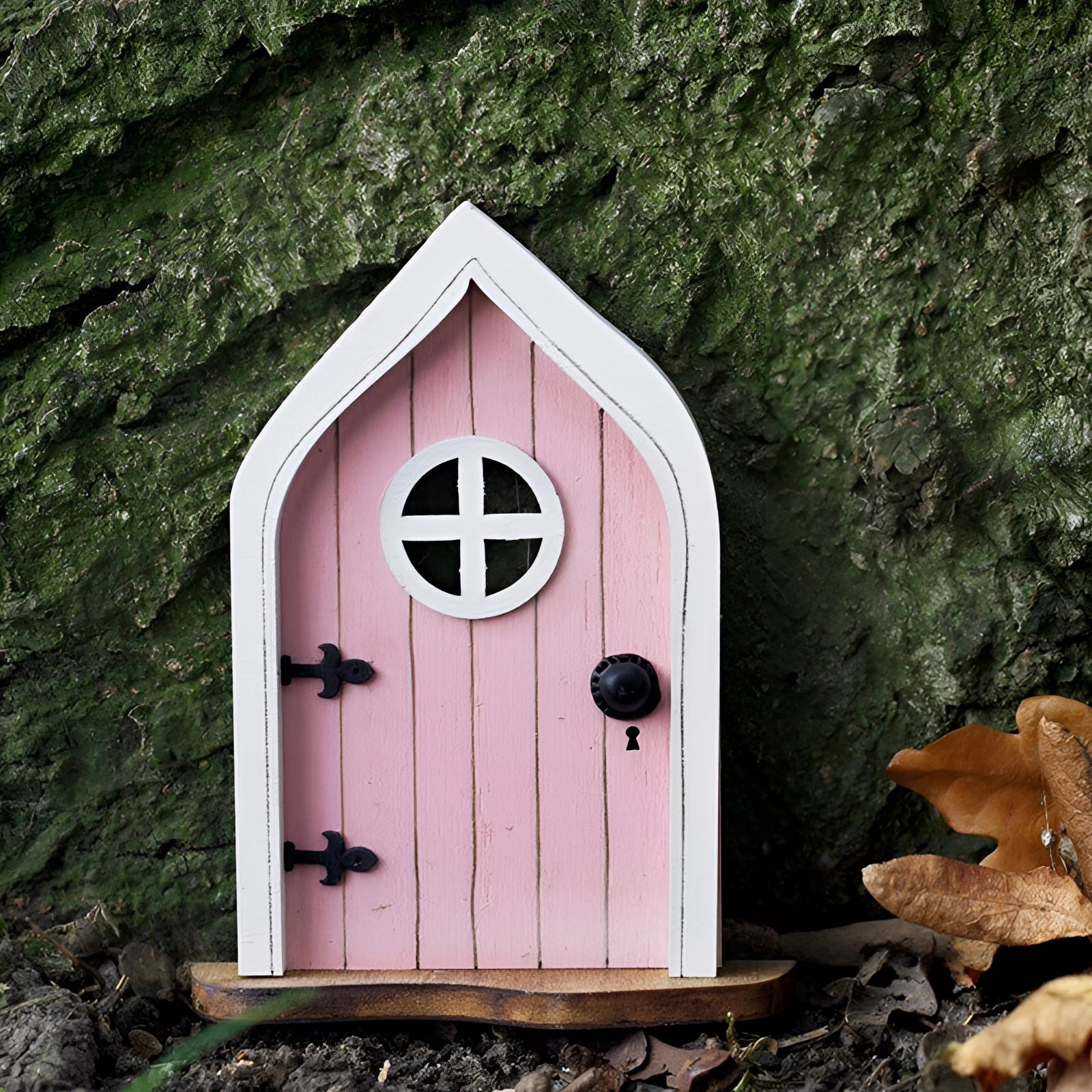 Miniature pink fairy garden door set within an enchanted tree, featuring a small window, surrounded by lush greenery and resembling a whimsical cottage with birdhouse accents.