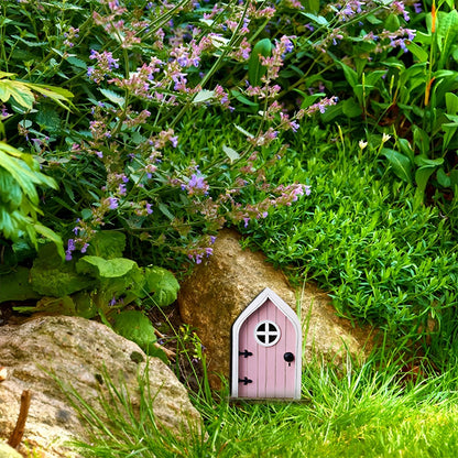 Miniature fairy garden door nestled in a lush green landscape with grass, shrubs, and small plants surrounding a whimsical, tree-like structure.